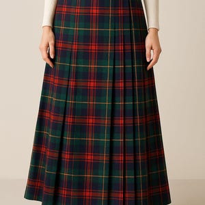 Puede incluir: Falda larga plisada a cuadros en tonos verdes, rojos y azul marino. La falda tiene una cintura alta con dos hebillas decorativas. La modelo lleva un top crema de manga larga y tacones negros.