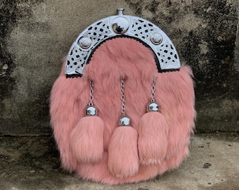 May include: A pink fur sporran with a silver cantle featuring Celtic knotwork and three matching tassels. The sporran has a rounded shape and a black trim. The background is a textured, neutral-toned surface.