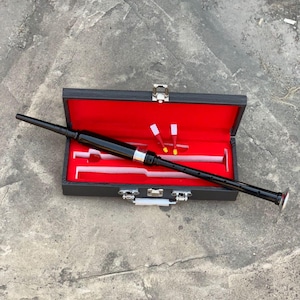 Puede incluir: Una gaita negra con detalles plateados, presentada en un estuche abierto con interior rojo. La gaita es negra con plata y un extremo blanco. El estuche es negro con cierres plateados.