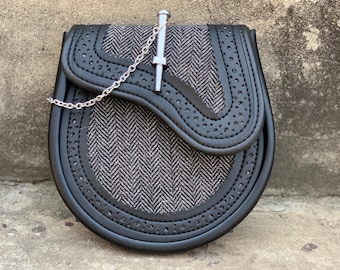 Puede incluir: Sporran de cuero negro con un patrón de espiga gris y blanco. El sporran tiene una cadena plateada y un alfiler decorativo plateado. Los bordes están perforados con pequeños agujeros.