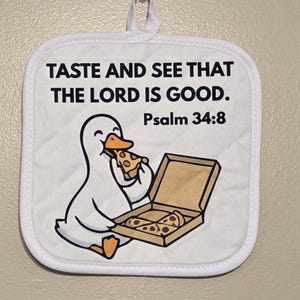 May include: White square pot holder with a cartoon duck eating pizza from a box. Text reads "TASTE AND SEE THAT THE LORD IS GOOD. Psalm 34:8". The pot holder has a white border and a loop for hanging.