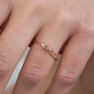 May include: A delicate gold ring featuring a knot design, worn on a finger. The ring has a slender band and a small, subtle knot detail. The ring is a simple yet elegant piece of jewellery.