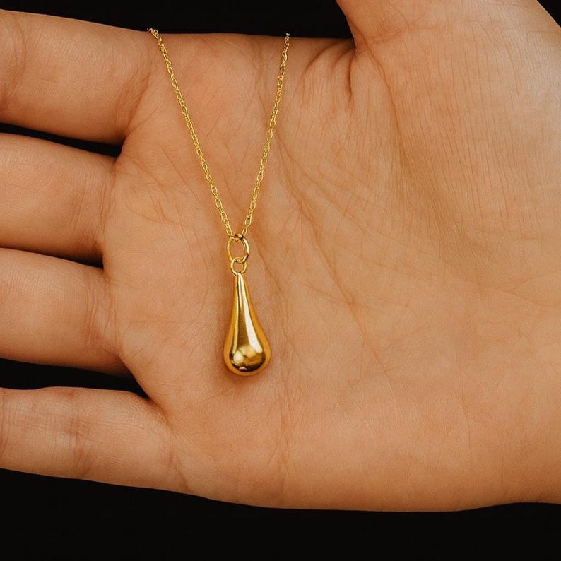 May include: A gold-colored teardrop pendant necklace. The pendant is a smooth, polished teardrop shape suspended from a delicate gold chain. The necklace is displayed against a neutral background.