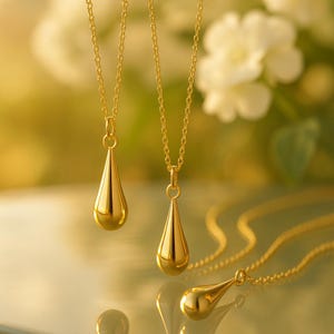 May include: Several gold-colored teardrop pendant necklaces with delicate chains. The pendants have a smooth, polished finish, reflecting the light. The necklaces are displayed against a soft, blurred background, highlighting the jewelry's elegant design.