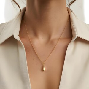 May include: A gold-colored pendant necklace with a teardrop-shaped charm. The necklace is worn against a cream-colored collared shirt. The pendant hangs from a delicate chain, creating a simple and elegant look. The jewelry is a classic accessory.