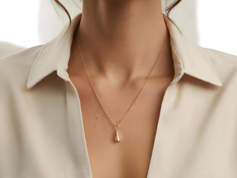 May include: A gold-colored pendant necklace with a teardrop-shaped charm. The necklace is worn against a cream-colored collared shirt. The pendant hangs from a delicate chain, creating a simple and elegant look. The jewelry is a classic accessory.