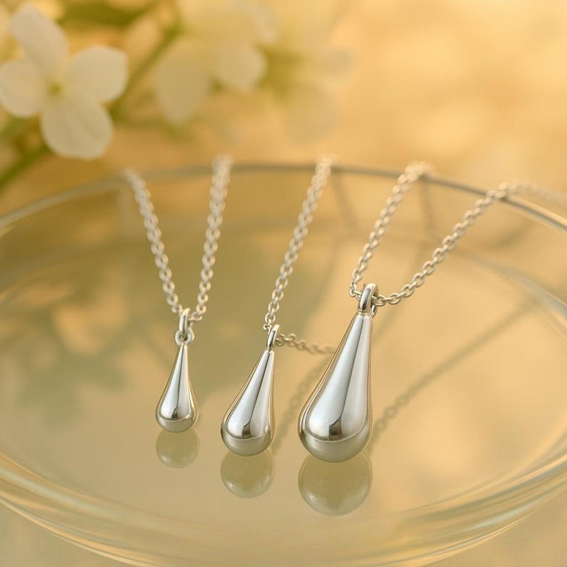 May include: Three silver teardrop pendant necklaces of varying sizes. Each pendant hangs from a delicate silver chain. The pendants are smooth and reflective, with a polished finish. The necklaces are displayed on a reflective surface.