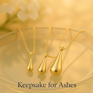 May include: Three gold-colored teardrop-shaped pendants on delicate chains. The pendants vary in size, with the largest one in the foreground. The text "Keepsake for Ashes" is at the bottom of the image.