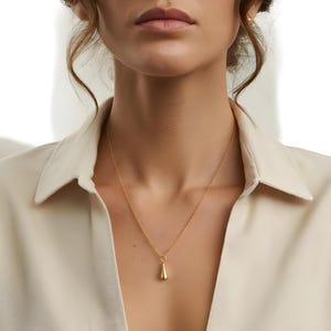 May include: A gold-colored pendant necklace with a teardrop-shaped charm. The necklace is worn against a cream-colored collared shirt. The pendant hangs from a delicate chain, creating a simple and elegant look.