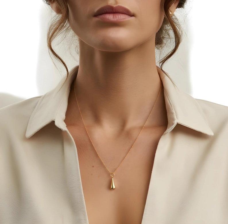 May include: A gold-colored pendant necklace with a teardrop-shaped charm. The necklace is worn against a cream-colored collared shirt. The pendant hangs from a delicate chain, creating a simple and elegant look.