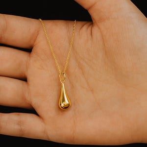 May include: A gold-colored pendant necklace featuring a teardrop-shaped charm. The charm is suspended from a delicate chain. The necklace is displayed against a neutral background.
