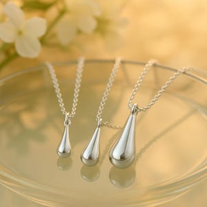 May include: Three silver teardrop pendant necklaces of varying sizes. Each pendant hangs from a delicate silver chain. The pendants are smooth and reflective, with a polished finish. The necklaces are displayed on a reflective surface.