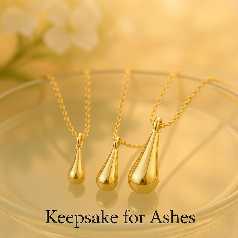 May include: Three gold-colored teardrop-shaped pendants on delicate chains. The pendants vary in size, with the largest one in the foreground. The text "Keepsake for Ashes" is at the bottom of the image.
