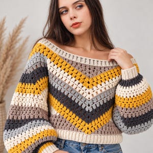 May include: A long-sleeved crochet sweater featuring a chevron pattern in horizontal stripes of mustard yellow, cream, light gray, and dark charcoal gray. The garment has a wide boat neckline and ribbed cuffs and hem in cream.
