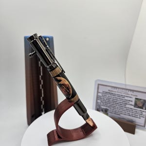 Könnte beinhalten: Ein handgefertigter Stift mit einem schwarzen und holzmaserierten Schaft, der ein dekoratives Design aufweist. Der Stift wird auf einem roten Ständer präsentiert, mit einem dunkelbraunen Ständer im Hintergrund. Ein Zertifikat ist im Hintergrund sichtbar.