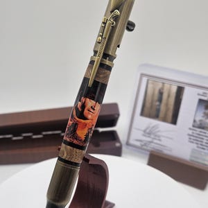 Peut inclure: Un stylo unique avec un design en forme de balle de couleur bronze. Le stylo présente une image détaillée d'une personne et d'un clip de fusil. Le stylo est exposé sur un support en bois, avec une boîte en bois et un certificat en arrière-plan.