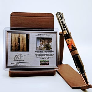 Peut inclure: Un stylo artisanal sur le thème de John Wayne, présenté sur un présentoir en bois. Le stylo présente une image détaillée de John Wayne et un corps en grain de bois. Un certificat d'authenticité est affiché derrière le stylo.