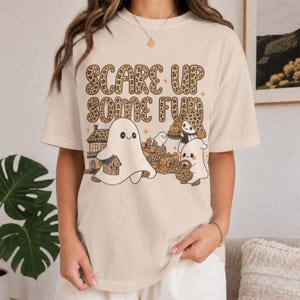 Op de afbeelding: Een crèmekleurig t-shirt met de tekst "SCARE UP SOME FUN" in een luipaardprint. De afbeelding bevat drie cartoonspoken, een huis en pompoenen, allemaal uitgevoerd in een bruin en oranje kleurenpalet met kleine steraccenten.