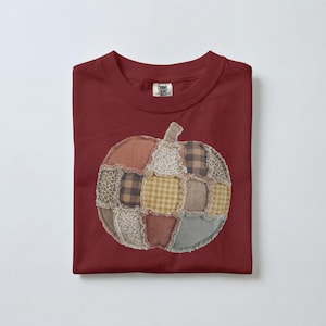 May include: A dark red crew neck t-shirt featuring a central pumpkin graphic composed of various fabric patches. The pumpkin design includes squares of plaid, gingham, floral, leopard print, and solid textures with frayed edges.
