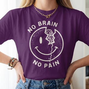 May include: A dark purple t-shirt featuring a white graphic of a cracked smiley face with a brain visible in the crack. The text NO BRAIN NO PAIN is printed in white in a circular arc around the smiley face design on the front of the shirt.
