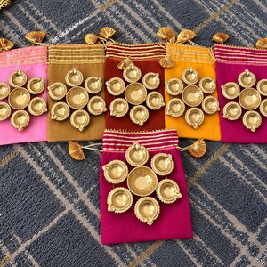 May include: Six rectangular fabric drawstring pouches in pink, tan, maroon, yellow, and magenta. Each pouch features a gold-toned metallic floral arrangement of seven small oil lamps, with striped fabric tops and gold tassel accents.