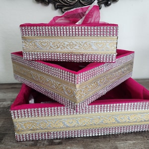 May include: Three stacked decorative boxes in varying sizes. The boxes are a vibrant magenta color, with gold and silver patterned trim. The trim is embellished with small, sparkling rhinestones. The boxes appear to be made of fabric and are nested together.