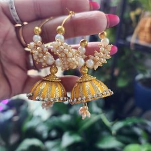 May include: Gold hoop earrings with yellow and gold striped bell-shaped dangles. The earrings are adorned with small white pearls and clear rhinestones. The earrings are a unique and eye-catching accessory.