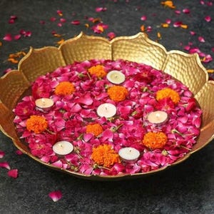 May include: A gold-toned metal bowl with a scalloped lotus-shaped rim filled with water, scattered pink rose petals, bright orange marigold flowers, and several small white tea light candles floating on the surface of the water.