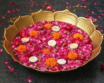 May include: A gold-toned metal bowl with a scalloped lotus-shaped rim filled with water, scattered pink rose petals, bright orange marigold flowers, and several small white tea light candles floating on the surface of the water.