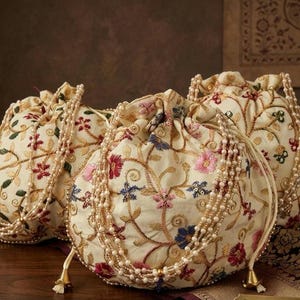 May include: Three off-white drawstring pouches featuring intricate floral embroidery in shades of red, pink, blue, and green. Each bag is adorned with multiple strands of white faux pearls that serve as handles and decorative accents with gold-toned tassels.