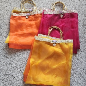 May include: Three sheer fabric tote bags in shades of orange, yellow, and pink. Each bag has a gold handle and a decorative trim along the top edge. A small floral embellishment hangs from the center of the trim.