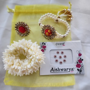 May include: A collection of Indian jewelry and accessories. Includes a pearl bracelet with a decorative centerpiece, matching earrings, a floral garland, and a packet of bindi stickers. The items are arranged on a yellow fabric.