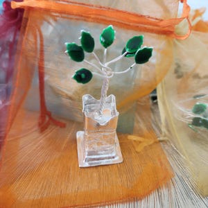 May include: A small, silver-colored metal tree sculpture with green enamel leaves. The tree is set in a square base with a small candle holder. The background includes orange and yellow sheer fabric.