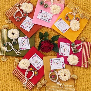 May include: Six fabric pouches in orange, pink, yellow, green, red, and gold, each featuring gold trim, a white floral accessory, a beaded bracelet, and a pack of Aishwarya bindis. A single red rose is positioned in the center.