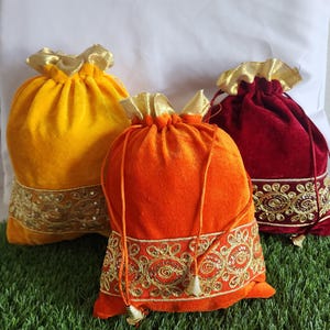 May include: Three velvet drawstring bags in shades of yellow, orange, and burgundy. Each bag features a gold-colored, sequined trim along the bottom and a gold-colored drawstring closure. The bags are on a green surface.