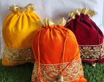 May include: Three velvet drawstring bags in shades of yellow, orange, and burgundy. Each bag features a gold-colored, sequined trim along the bottom and a gold-colored drawstring closure. The bags are on a green surface.