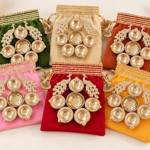 May include: Six velvet drawstring pouches in green, beige, orange, pink, red, and yellow, each featuring a metallic gold-toned peacock-shaped diya lamp holder with seven circular candle cups arranged in a circular pattern with decorative tassels.