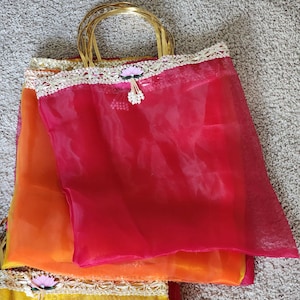 May include: A stack of colorful, sheer tote bags with gold handles. The bags are in shades of red, orange, and yellow, with decorative lace trim and floral accents. The bags are likely for gifts or special occasions.