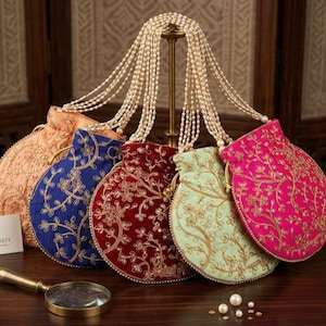 May include: Five round drawstring pouches in peach, blue, dark red, light green, and fuchsia, each featuring gold floral embroidery and a long pearl bead handle. The pouches are displayed on a stand with a magnifying glass and business card.