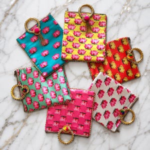 May include: Six square fabric pouches with circular gold-toned metal handles, each featuring a repeating lotus flower pattern. The pouches come in teal, yellow, red, light green, hot pink, and white, with small decorative tassels attached to the handles.