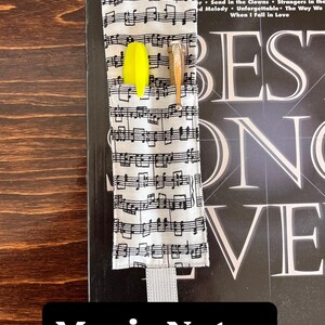 Puede incluir: Un marcap&aacute;ginas de tela con un patr&oacute;n de notas musicales, con un resaltador amarillo y un l&aacute;piz de madera. El marcap&aacute;ginas est&aacute; colocado en un libro titulado "The Best Songs Ever". El texto "Music Notes" se muestra en un rect&aacute;ngulo negro.