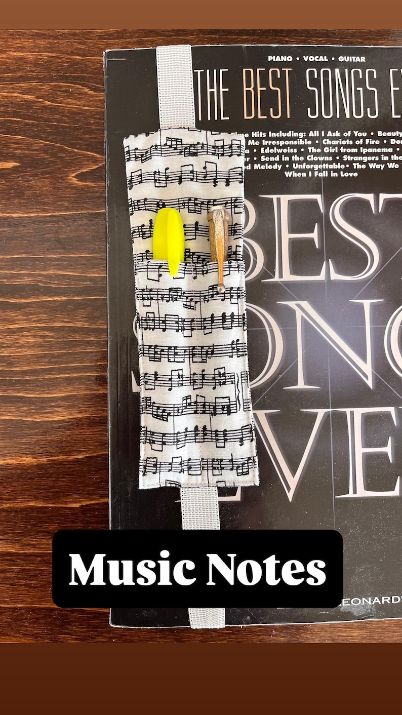 Puede incluir: Un marcap&aacute;ginas de tela con un patr&oacute;n de notas musicales, con un resaltador amarillo y un l&aacute;piz de madera. El marcap&aacute;ginas est&aacute; colocado en un libro titulado "The Best Songs Ever". El texto "Music Notes" se muestra en un rect&aacute;ngulo negro.