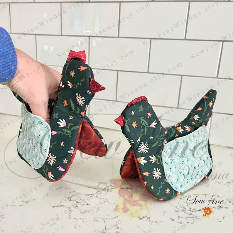 May include: Two chicken-shaped oven mitts. The mitts are dark green with a floral pattern and red accents. The interior is red, and the mitts have a light blue quilted section. One mitt is being held by a person.
