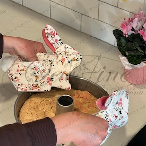 Peut inclure: Des maniques en forme de lapin avec des motifs floraux sont utilis&eacute;es pour manipuler un g&acirc;teau. Les mitaines ont une doublure rose et des queues en pompon blanc. Le g&acirc;teau est dans un moule rond.