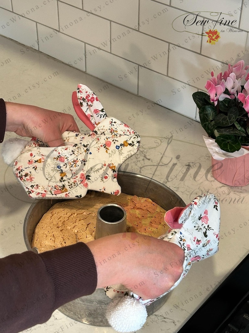 Peut inclure: Des maniques en forme de lapin avec des motifs floraux sont utilis&eacute;es pour manipuler un g&acirc;teau. Les mitaines ont une doublure rose et des queues en pompon blanc. Le g&acirc;teau est dans un moule rond.