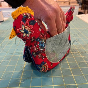 May include: A handmade fabric chicken-shaped item, primarily red with a floral pattern, blue accents, and a yellow comb and beak. The chicken has a pocket on its side. The item is likely a kitchen accessory or decorative piece.