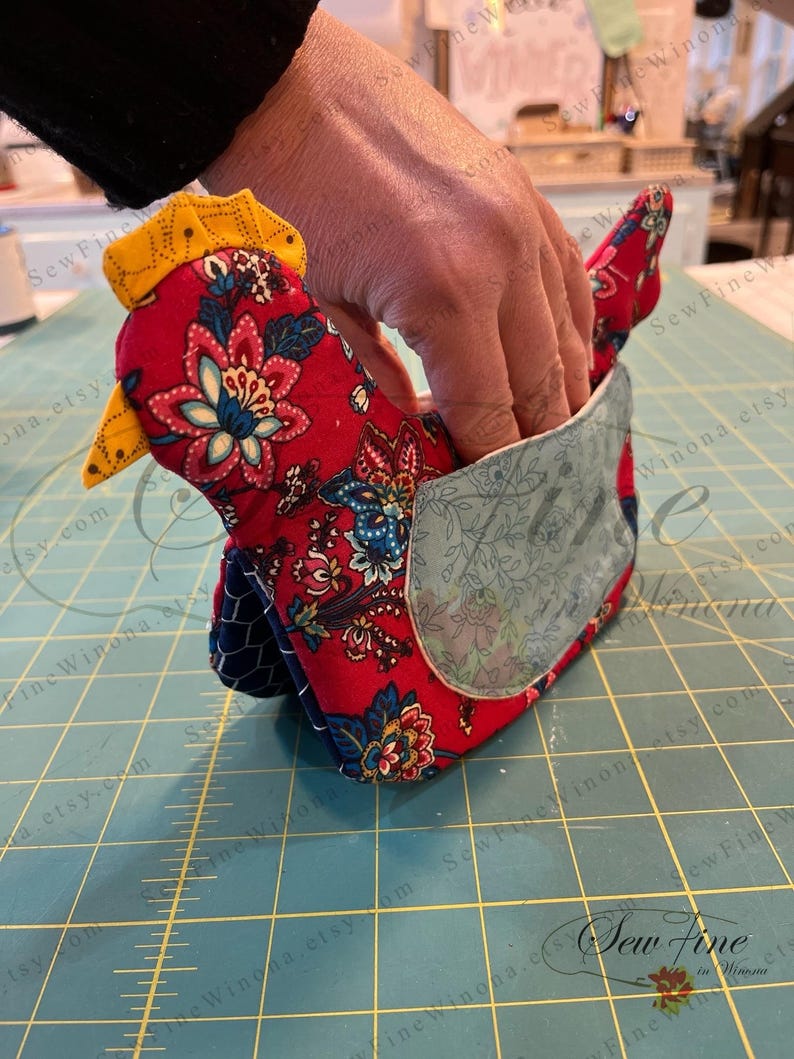 May include: A handmade fabric chicken-shaped item, primarily red with a floral pattern, blue accents, and a yellow comb and beak. The chicken has a pocket on its side. The item is likely a kitchen accessory or decorative piece.