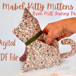 May include: A cat-shaped oven mitt sewing pattern. The mitt is made of fabric with a floral pattern in red, brown, and cream colors. The mitt has a green collar and a tail. The text "Mabel Kitty Mittens Oven Mitt Sewing Pattern" is visible.
