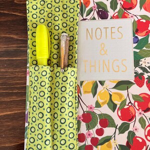 Puede incluir: Cuaderno con un colorido estampado de frutas, con limones, cerezas y manzanas. Tiene un bolsillo verde con un dise&ntilde;o de c&iacute;rculos que contiene un resaltador amarillo y un bol&iacute;grafo. El texto "Spring Green" y "NOTES & THINGS" son visibles.