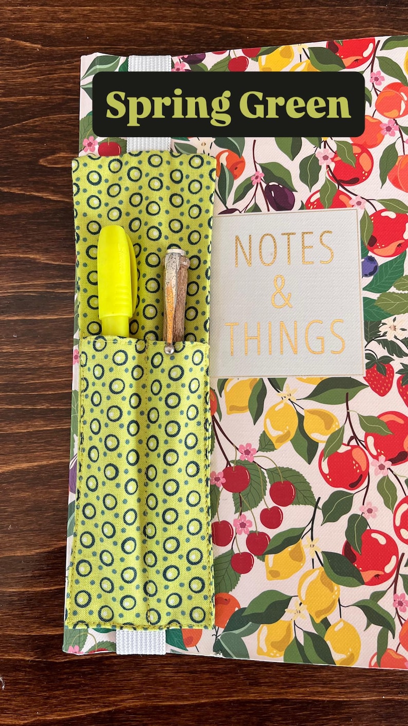 Puede incluir: Cuaderno con un colorido estampado de frutas, con limones, cerezas y manzanas. Tiene un bolsillo verde con un dise&ntilde;o de c&iacute;rculos que contiene un resaltador amarillo y un bol&iacute;grafo. El texto "Spring Green" y "NOTES & THINGS" son visibles.